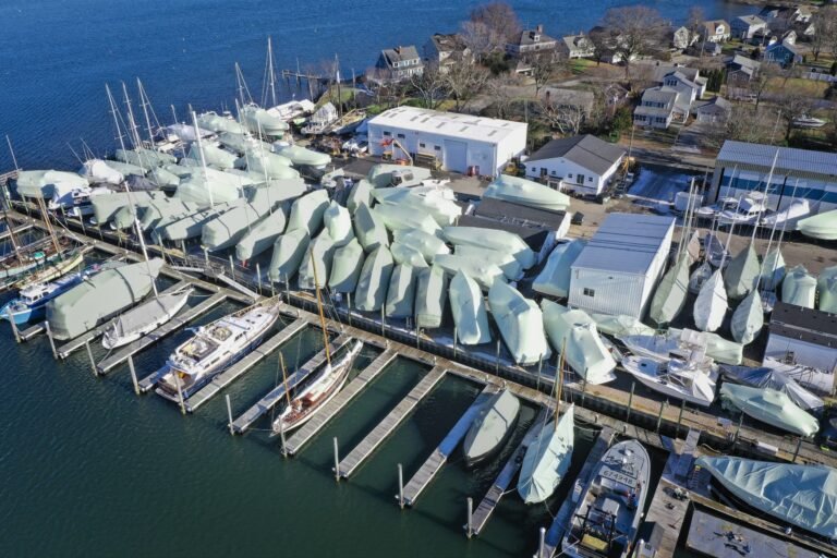 bioaqualife: Aerial view of a marina with many boats covered in white protective tarps, docked at piers near waterfront buildings and houses, with water and trees in the background.