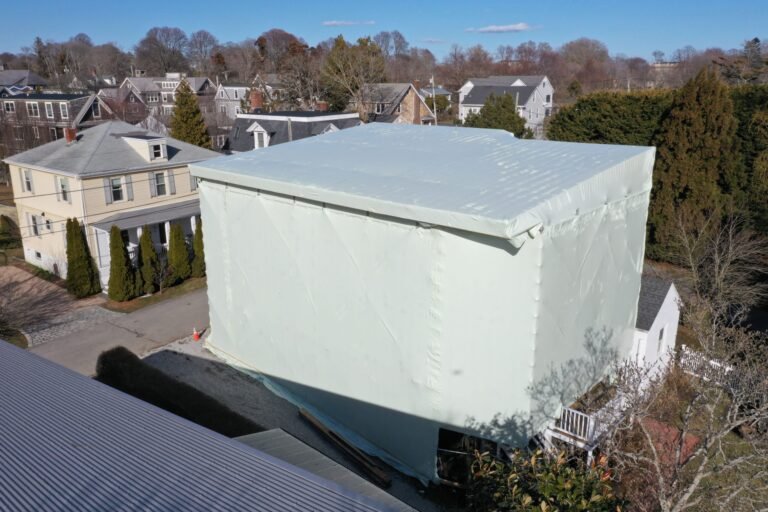 bioaqualife: A large house covered entirely in white plastic sheeting, likely for renovation, stands in a residential neighborhood with other houses and trees visible in the background under a clear sky.