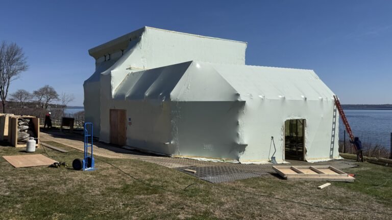bioaqualife: A large building near a body of water is covered in white plastic sheeting. A ladder leans against one side, and construction materials and equipment are scattered on the grass around it under a clear sky.