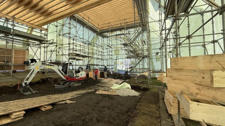 bioaqualife: Interior view of a construction site with scaffolding, a small excavator, wooden planks, gravel, and unfinished flooring. Sunlight filters through translucent plastic sheeting along the walls and roof.