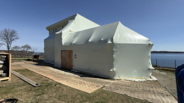 bioaqualife: A small building near a body of water is completely covered in white plastic sheeting, with a wooden door and signs visible on one side. Wooden planks and construction materials are scattered on the ground nearby.