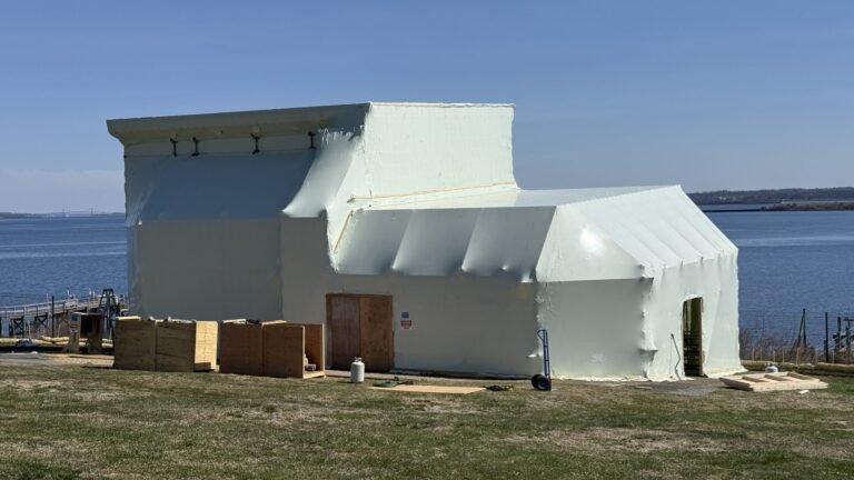 bioaqualife: A large, oddly shaped building is covered in pale insulation material near a body of water; wooden crates and construction materials are scattered on the grass around it under a clear blue sky.