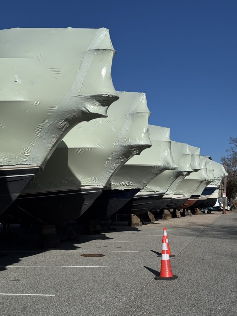 bioaqualife: A row of boats covered in pale green protective shrink wrap are elevated on blocks in a parking lot, with a single orange traffic cone in the foreground and a clear blue sky overhead.