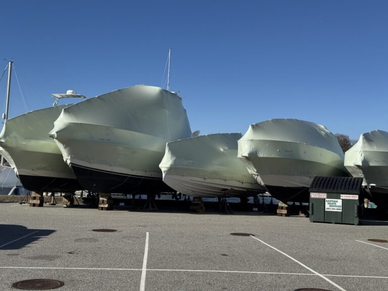 bioaqualife: Several large boats covered in protective shrink wrap are stored on blocks in a parking lot under a clear blue sky. A green dumpster is nearby, and parking lines are visible on the ground.