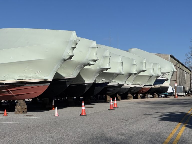 bioaqualife: Several large boats covered in white shrink wrap are lined up on blocks in a parking lot, with orange traffic cones placed around them under a clear blue sky.