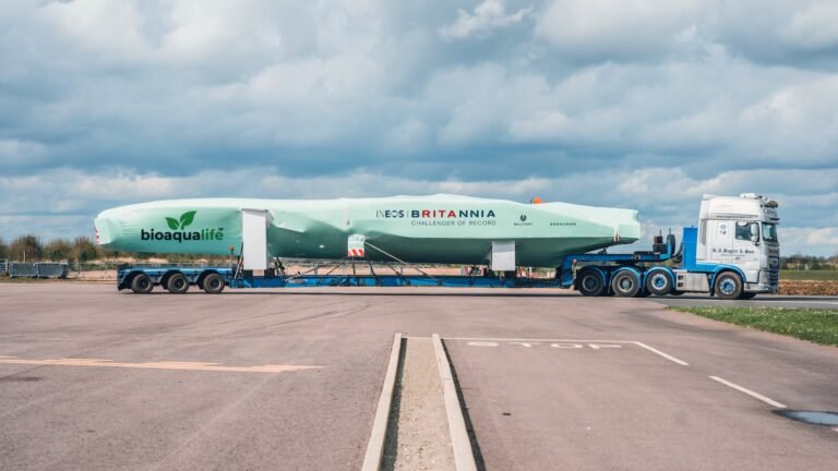 bioaqualife: A long, green boat hull labeled "INEOS Britannia" and "bioqualif&eacute;" is transported on a flatbed trailer pulled by a white truck on a paved road under a cloudy sky.