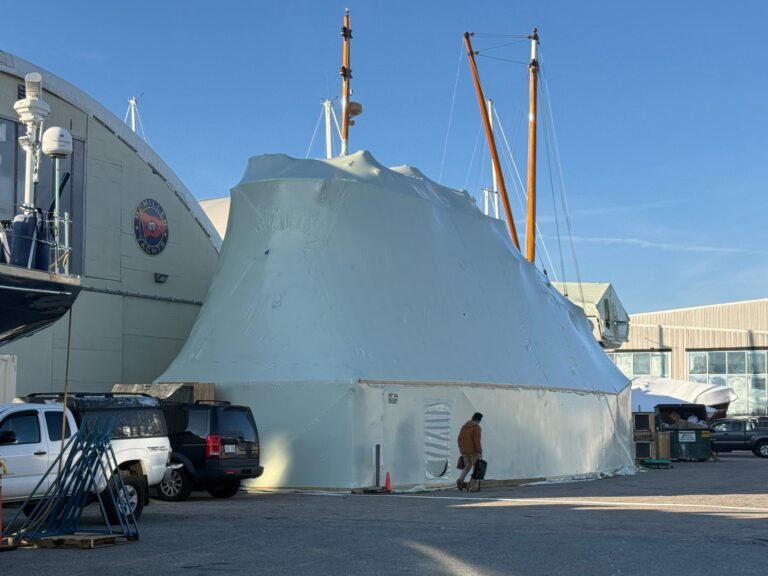 bioaqualife: A large boat is covered in white shrink wrap at a shipyard, with parked vehicles nearby and a person walking past. Mast poles extend above the wrapped boat, and industrial buildings are in the background under a clear blue sky.