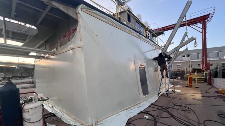 bioaqualife: A person stands on a ladder working on the side of a large boat wrapped in protective plastic at a shipyard, with tools, hoses, and equipment scattered around. The evening sky is visible in the background.