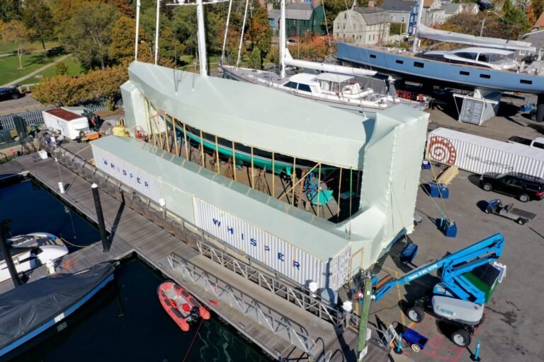 bioaqualife: A large sailboat is under construction or repair at a dock, covered with a white canopy and flanked by shipping containers labeled &ldquo;WHISPER.&rdquo; Construction equipment and boats are nearby, with trees and buildings in the background.