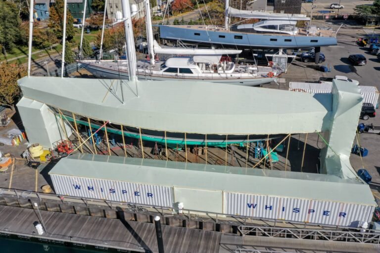 bioaqualife: A large boat is covered with a pale green temporary structure at a dockyard, surrounded by scaffolding and shipping containers labeled "WHISPER." Other yachts and buildings are visible in the background.