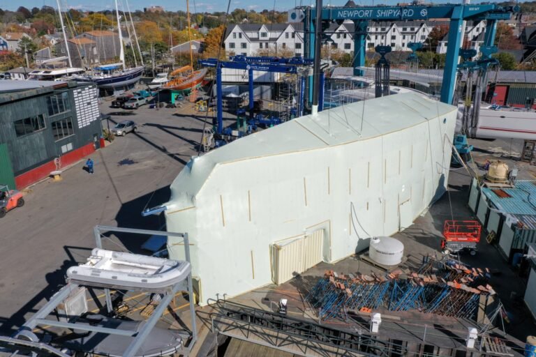 bioaqualife: A large boat hull covered in white protective material sits in a busy shipyard with cranes, equipment, boats, and buildings visible around it on a clear day.