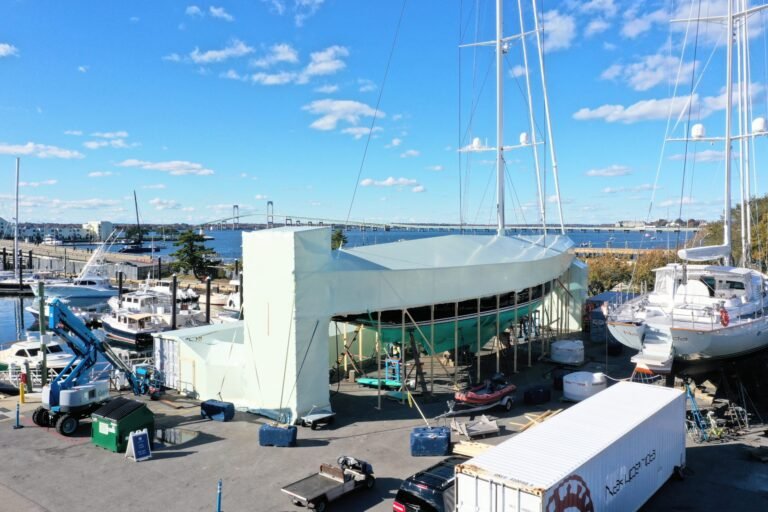 bioaqualife: A large sailboat is covered with a white tarp for repairs at a marina. Other boats and equipment are nearby, with a bridge and blue water visible in the background under a partly cloudy sky.