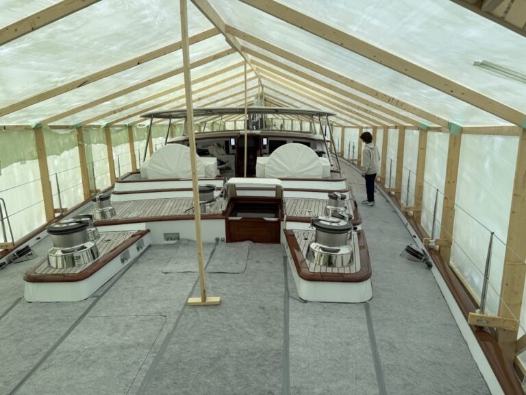 bioaqualife: A person stands near the side of a large yacht covered by a temporary wooden and plastic shelter. The deck is clean, with covered seating areas and exposed winches visible under the transparent roof.