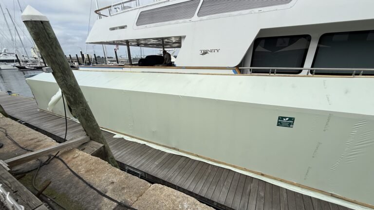 bioaqualife: A large boat docked at a marina, with its side covered by a long pale green protective barrier. The dock is wooden, and several boats and masts are visible in the background under a cloudy sky.
