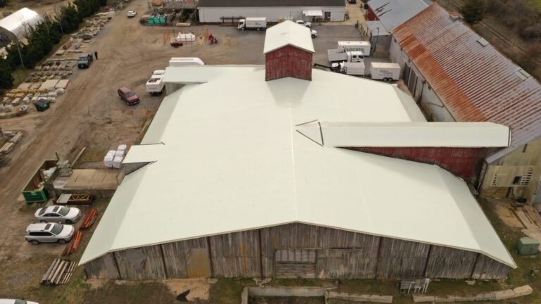 bioaqualife: Aerial view of a large, rustic, weathered wooden barn with a white roof, surrounded by dirt paths, parked vehicles, and industrial equipment in a rural setting.