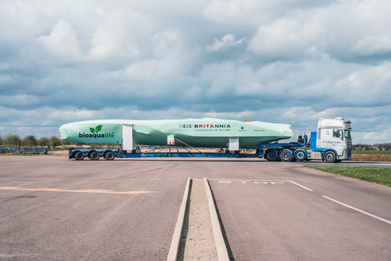 bioaqualife: A large truck transports a long, covered racing boat labeled "INEOS Britannia" and "bioaqualife" on a flatbed trailer across an empty paved area under a cloudy sky.