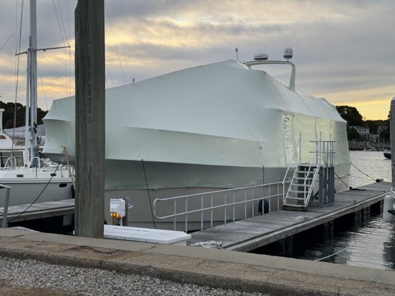 bioaqualife: A large boat covered in white shrink wrap is docked at a marina. There is a metal gangway attached to the boat and cloudy skies overhead. Other boats and houses are visible in the background.