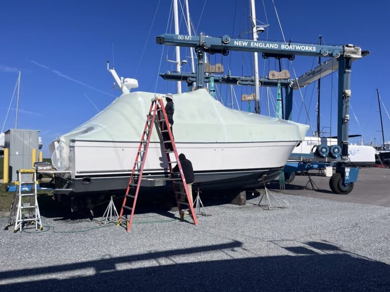 bioaqualife: A person stands on a ladder next to a white boat wrapped in a protective cover, supported by stands, with a boat lift labeled "New England Boatworks" and other boats visible in a marina background.