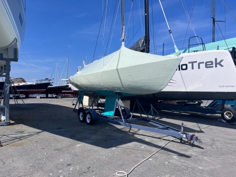 bioaqualife: A small sailboat covered in a light-colored protective wrap sits on a trailer in a boatyard, surrounded by other larger boats and sailboats under a clear blue sky.