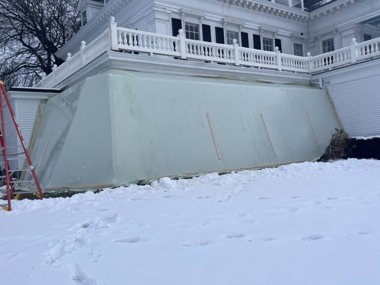 bioaqualife: A large, translucent plastic tarp covers part of a white house's porch in a snowy yard. Wooden boards and a ladder are visible nearby, suggesting ongoing construction or protection work.