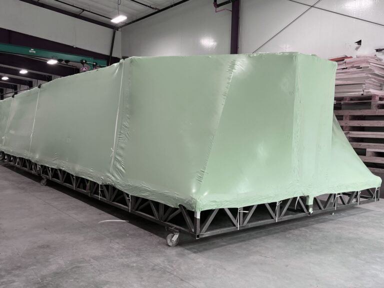 bioaqualife: A large, rectangular object covered with green plastic sits on a metal frame with wheels inside an industrial warehouse with white walls and concrete floors.