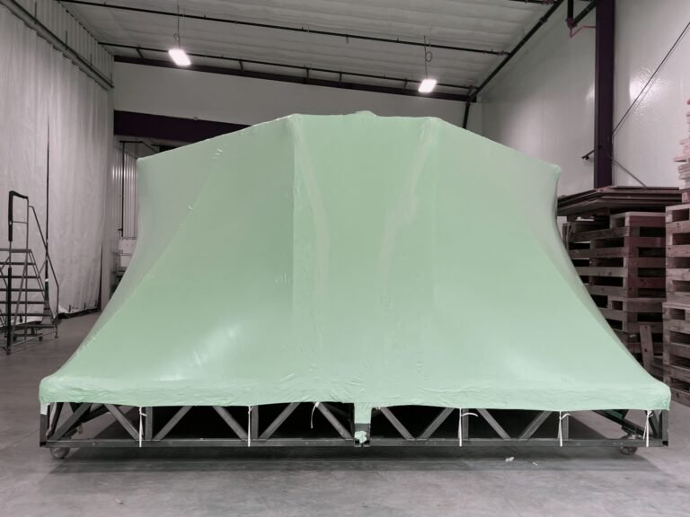 bioaqualife: A large, angular object is covered with smooth, green plastic sheeting in a spacious warehouse. The object rests on a metal frame, and stacks of wooden pallets are visible to the right.