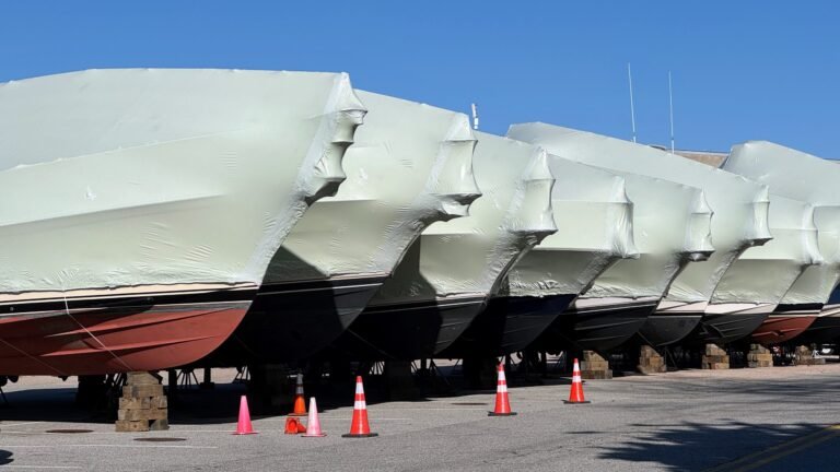 bioaqualife: Several boats are lined up on land, covered in protective white shrink wrap, elevated on blocks, with orange traffic cones placed in front of them. The sky is clear and blue.