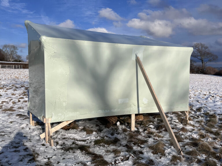 bioaqualife: A light green, rectangular shed on stilts stands in a snowy, grassy field. Wooden beams support one side, and the sky is partly cloudy with trees and a fence in the background.