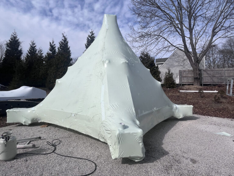 bioaqualife: A large, pale green structure covered tightly in plastic wrap stands outdoors on gravel, with trees and a house in the background under a partly cloudy sky.