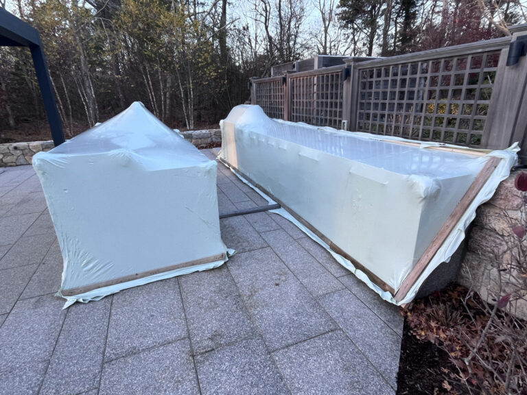 bioaqualife: Two large outdoor objects covered with light blue protective plastic wrap, sitting on a stone patio. Trees and wooden lattice fencing are visible in the background.