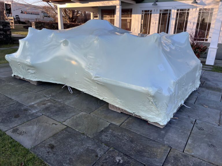 bioaqualife: A large object covered in white plastic is secured on pallets outdoors, in front of a house with a porch and stone patio. The plastic tightly wraps around the item, obscuring its shape and details.